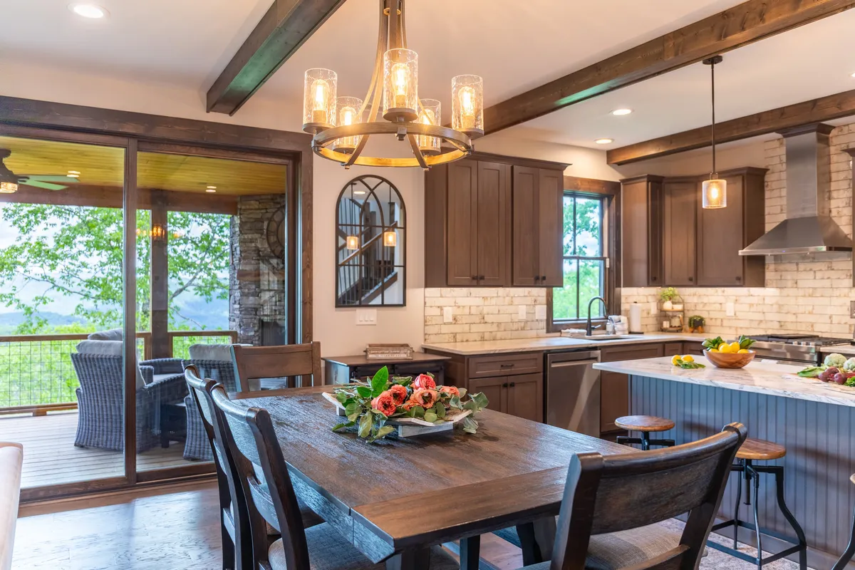 This image centers on a wooden dining table beneath a striking chandelier, situated beside a kitchen island and large windows. Decorative flowers provide a welcoming touch, and the generous windows frame scenic mountain vistas, bathing the room in natural light.