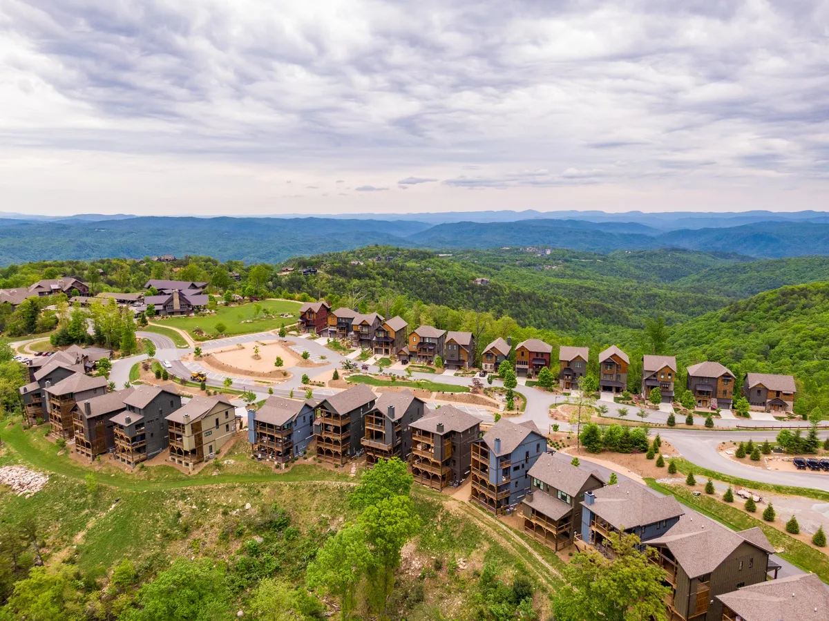 A wide-angle view of a mountainous landscape with residential buildings nestled against green hills and curved roadways. The area is surrounded by lush foliage, creating an inviting sense of outdoor adventure and connection to nature.