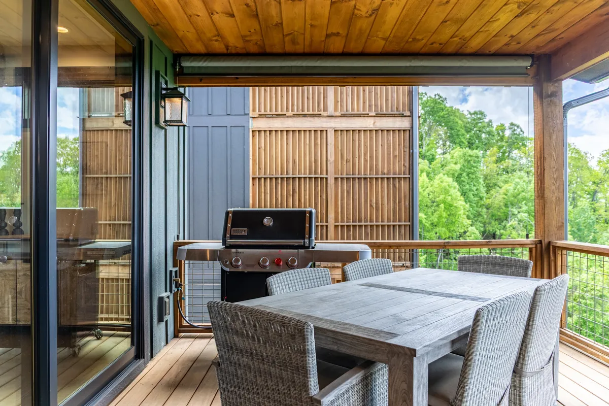 An exterior shot of a covered outdoor dining area featuring a wooden table, a barbecue grill, and a backdrop of tall green trees. The inviting setup suggests communal gatherings and summer cookouts.