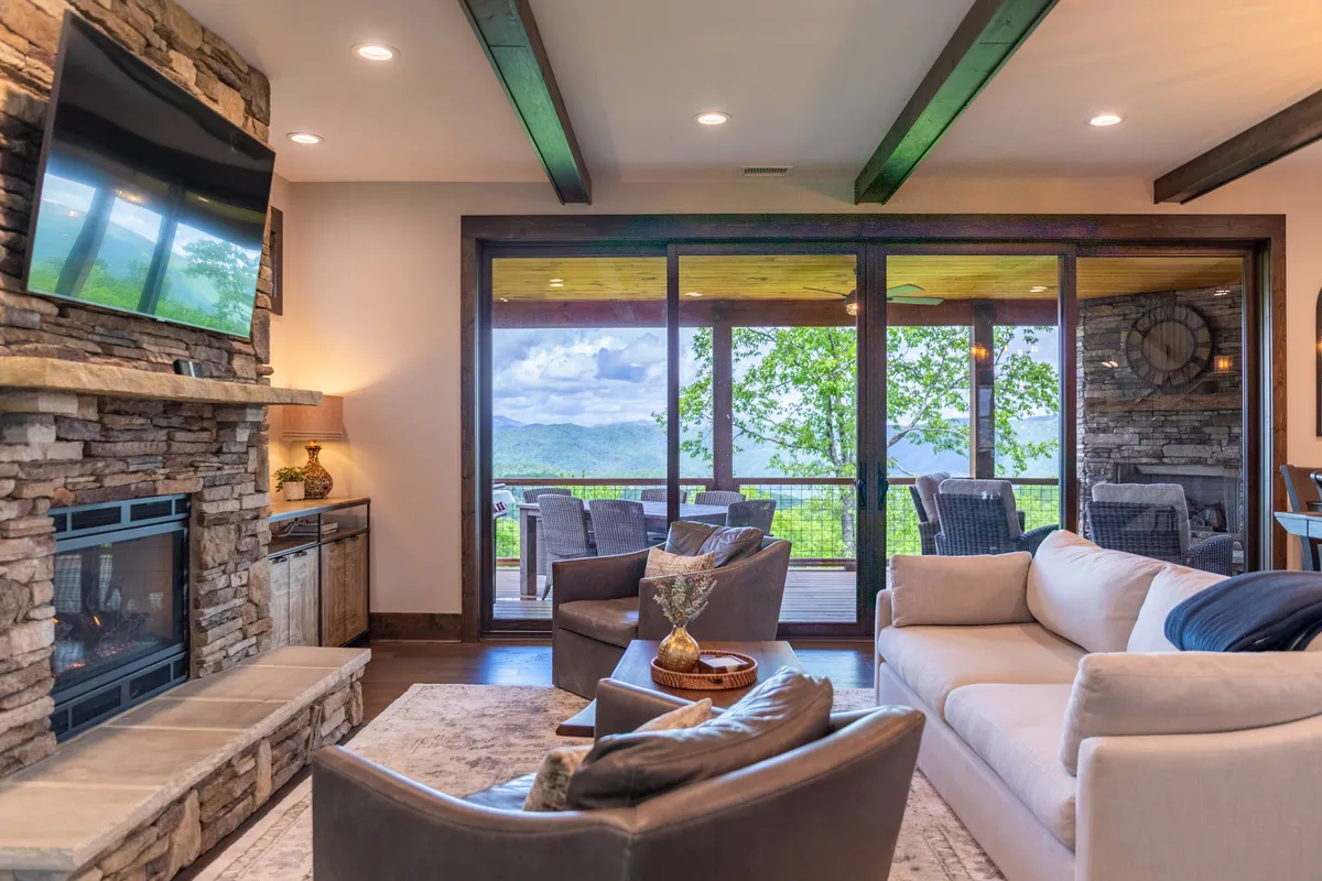 A bright, open living area featuring a beautiful stone fireplace, large windows framing expansive mountain views, and a plush, comfortable seating arrangement. Decorative accents and warm wood tones lend the space a contemporary mountain feel.