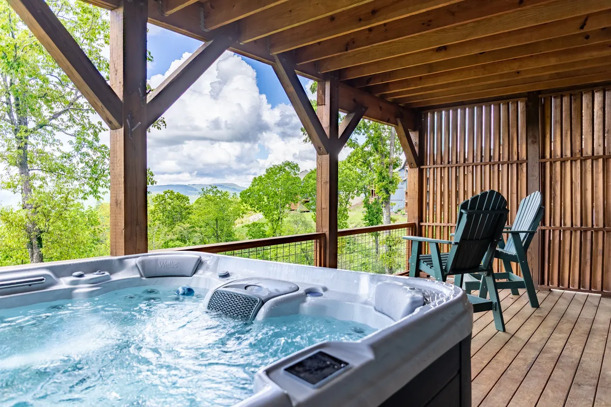 A serene outdoor space with a private hot tub and Adirondack chairs on a wooden deck, overlooking a peaceful view of trees and distant mountains. The setting is tranquil, offering sunset or early morning light, inviting relaxation and a strong connection to nature.