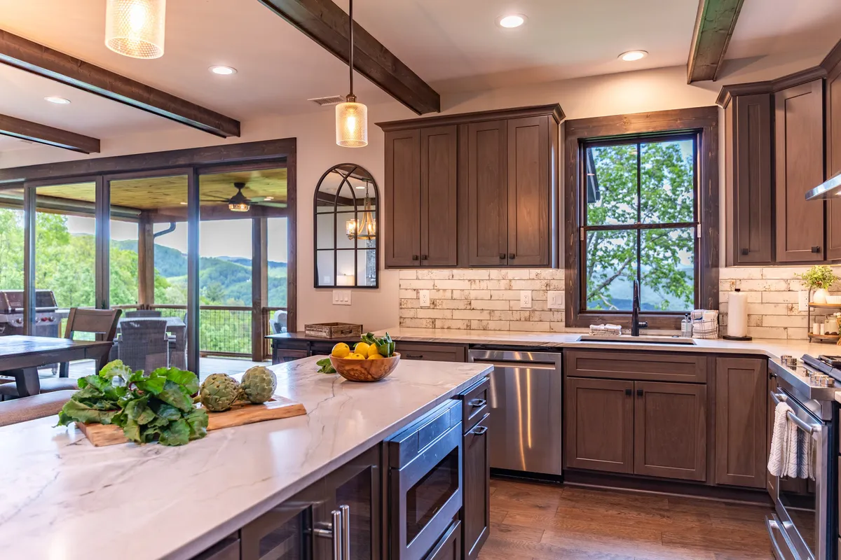 A welcoming kitchen island topped with fresh vegetables is the focal point, flanked by wooden cabinetry and beams. Large windows in the background invite natural light and offer a scenic view, creating a bright space ideal for cooking, entertaining, and family gatherings.