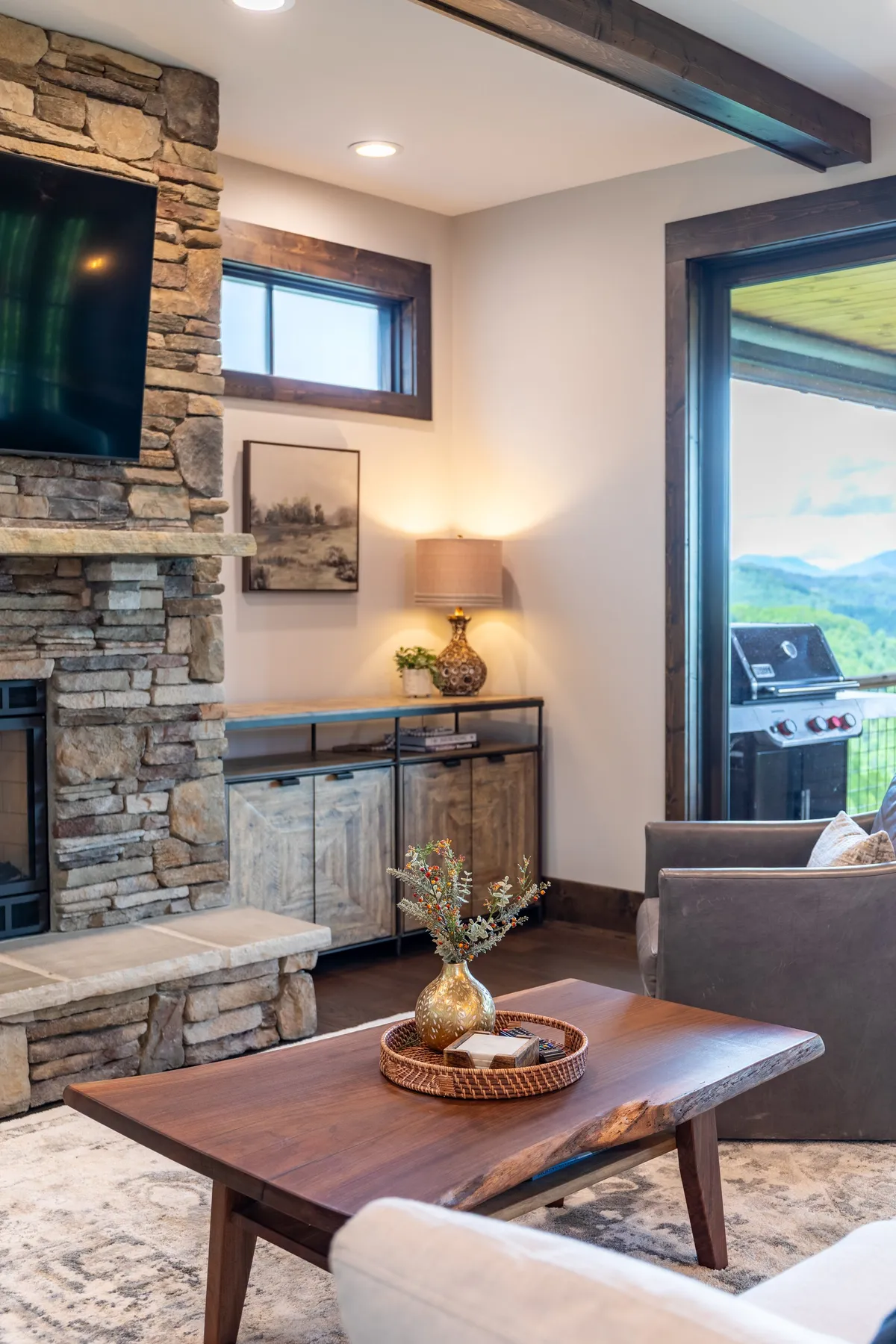 A welcoming living area with a stone fireplace, wooden coffee table, and a large window framing scenic mountain views. The room is bathed in natural light, suggesting warmth and inviting guests to gather and relax while enjoying the landscape outside.