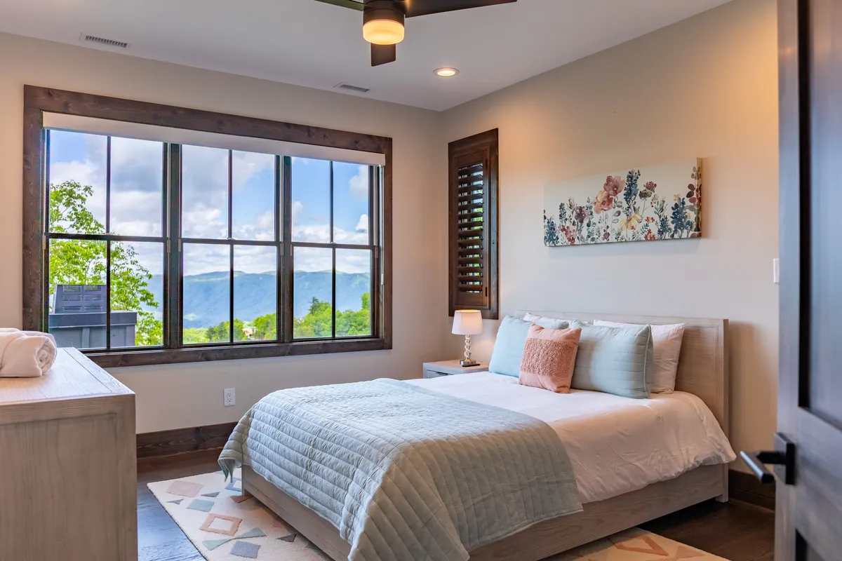 A cozy bedroom with a large window framing a stunning mountain view, a bed adorned with decorative pillows, and soft wall art. The peaceful setting is perfect for relaxing, reading, or simply enjoying the scenery from bed.