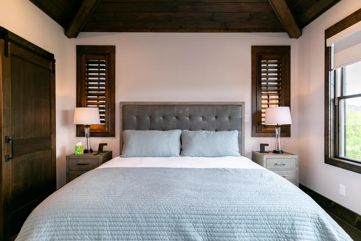 This image showcases a serene bedroom with a comfortable bed, nightstands, lamps, and a large window letting in abundant natural light. The scene exudes tranquility, inviting restful sleep and gentle morning wakeups.