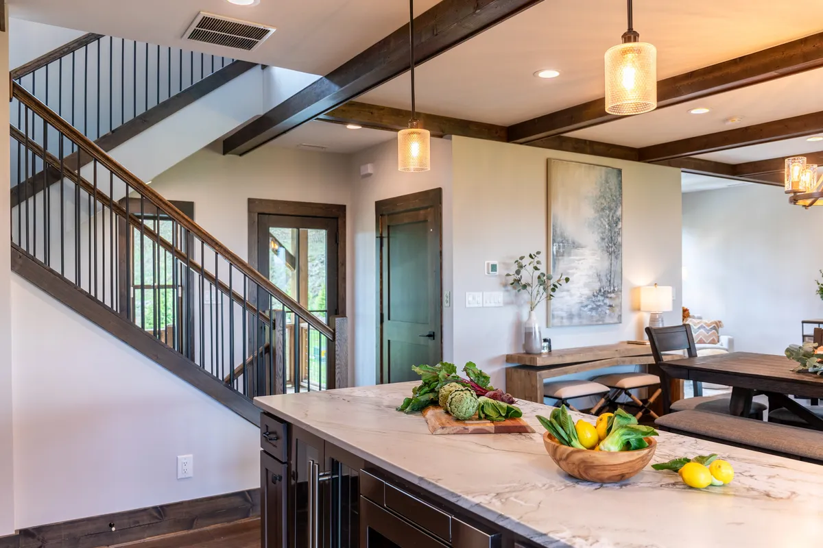 An open-plan space featuring a kitchen island, adjacent dining area, and stairs leading to other areas. Artwork and large windows add warmth and depth, and the layout encourages togetherness whether cooking, dining, or socializing.