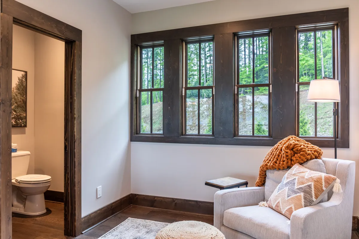 This image captures a comfortable chair positioned by large windows, offering a clear view of the outdoors. The scene suggests a tranquil moment of relaxing or reading while being visually connected to the surrounding landscape and the changing seasons visible through the window.