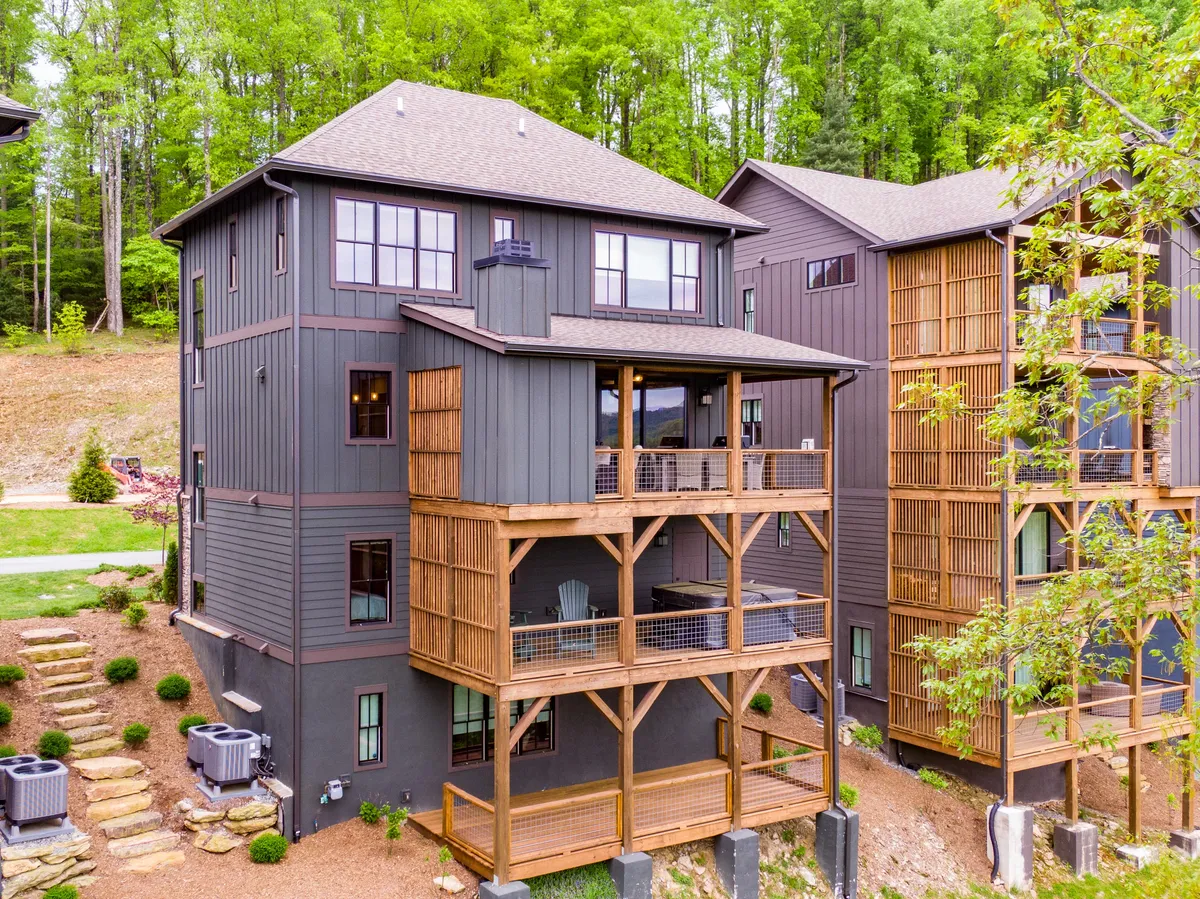 A multi-level cottage with dark wooden siding stands surrounded by green trees. Spacious wooden balconies extend outward, perfect for relaxing outdoors and enjoying sunrise or stargazing. The natural setting highlights the immersive mountain experience.