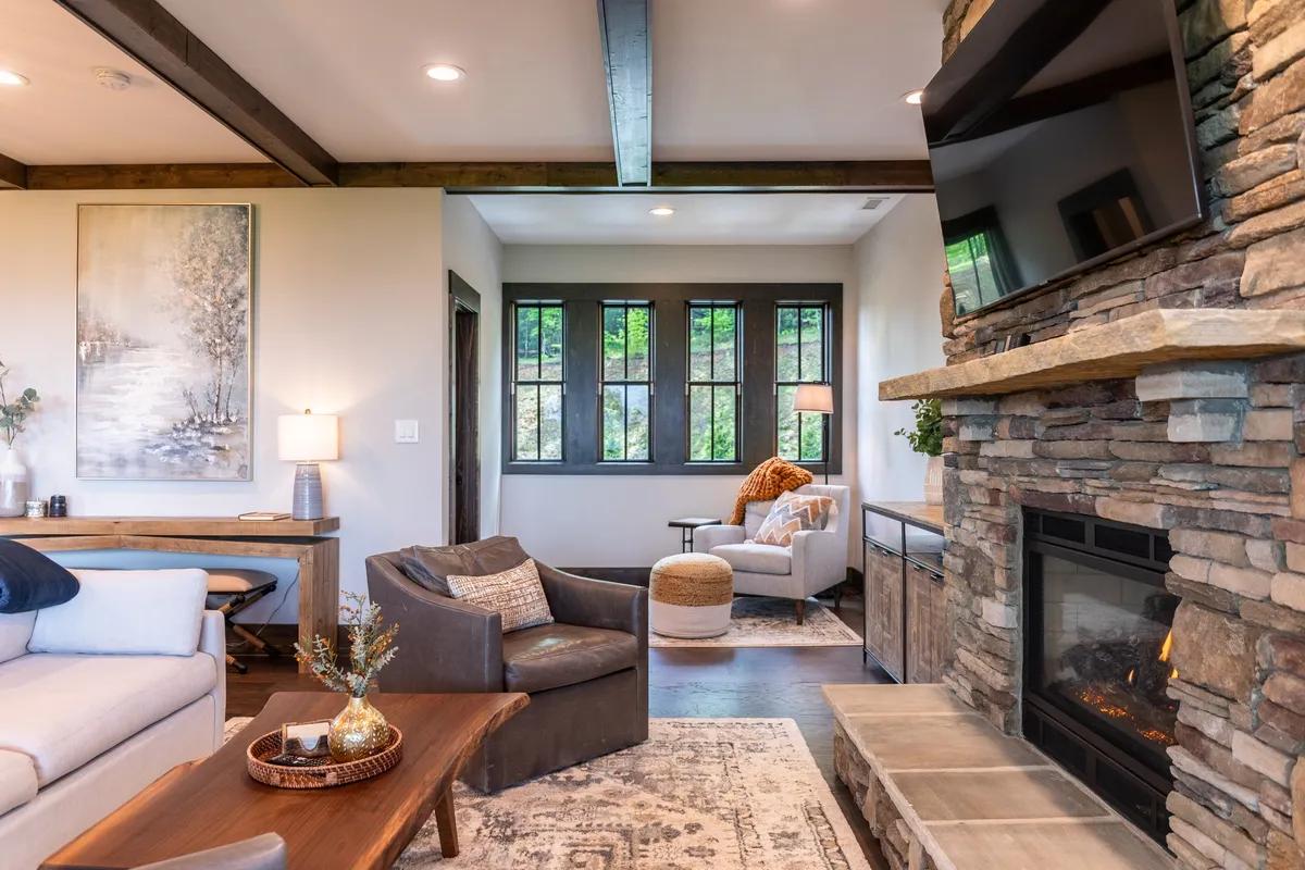 A cozy, inviting living room with a prominent stone fireplace, large windows flooding the space with natural light, and a welcoming seating arrangement. The room's décor and sunlight create a modern yet relaxed mountain vibe.